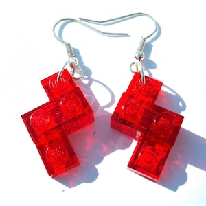 May include: A pair of red Lego earrings. Each earring is made of four red Lego bricks, connected together in a unique shape. The earrings are attached to silver hooks.