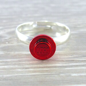 May include: A silver ring with a red LEGO brick design. The brick has the LEGO logo embossed on it.