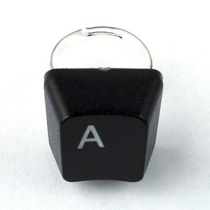 May include: A black keyboard key ring with a silver band. The key is shaped like a traditional computer key and has the letter "A" printed in white on it.