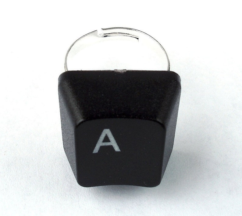 May include: A black keyboard key ring with a silver band. The key is shaped like a traditional computer key and has the letter "A" printed in white on it.