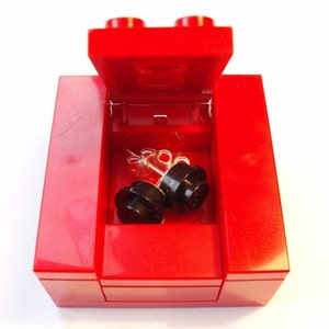 Puede incluir: Una caja roja de Lego con una tapa con bisagras, abierta para revelar un par de pendientes de botón negros con postes plateados.