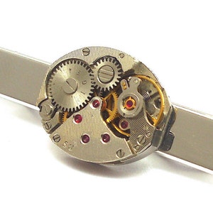 May include: A silver-toned tie clip featuring a watch movement. The watch mechanism is visible, showcasing gears, jewels, and other intricate components. The clip is rectangular and has a polished finish.