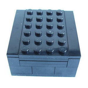 Puede incluir: Una caja de LEGO de color negro con una tapa que tiene 25 pequeños tacos negros en la parte superior. La caja está hecha de plástico y tiene una superficie lisa.