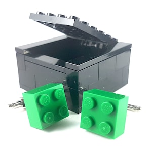 May include: A black LEGO brick box with a lid open, revealing two green LEGO brick cufflinks. The cufflinks are made of plastic and have the LEGO logo on each side.