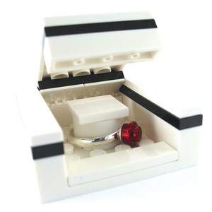 May include: A white Lego ring box with a black stripe on the lid. The lid is open and a silver ring is inside the box. There is a red Lego flower next to the ring.