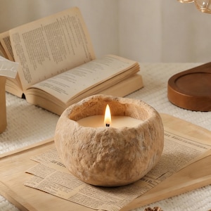 Op de afbeelding: Een brandende, ronde kaars in een getextureerde, beige container staat op een houten dienblad met krantenknipsels. Een open boek en een dennenappel zijn ook zichtbaar. De vlam van de kaars gloeit helder en creëert een warme sfeer.