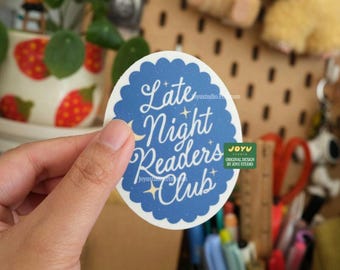 May include: A circular sticker featuring a blue scalloped edge and white text reading "Late Night Readers Club." The design includes stars and a crescent moon. The sticker is held in a hand.