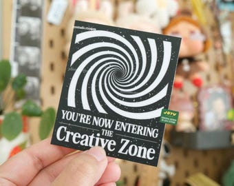 May include: Black and white sticker featuring a swirling spiral design. The text "You're Now Entering The Creative Zone" is printed on the sticker.