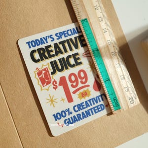 May include: A sticker with the text "TODAY'S SPECIAL CREATIVE JUICE" in black and blue. The sticker has a price of &pound;1.99 and guarantees 100% creativity. A wooden ruler with inch measurements is placed on the sticker.