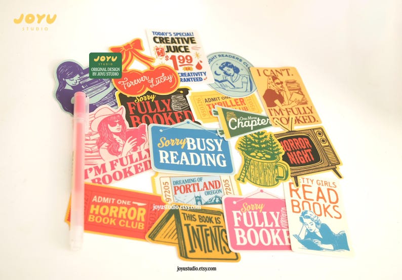 May include: A selection of colourful stickers featuring phrases like "Sorry Busy Reading" and "Horror Book Club," alongside illustrations. A pink pen is also visible. These stickers are perfect for book enthusiasts.