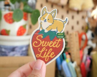 May include: A red and green strawberry sticker with a white outline. A cartoon corgi dog is sitting on top of the strawberry. The text "Sweet Life" is written in yellow on the strawberry.
