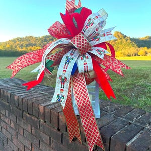 May include: A festive Christmas bow with red, white, and gold ribbons. The bow features a central design with a diamond pattern and ribbons with toy soldier illustrations. The bow is placed on a white lantern, perfect for holiday decor.
