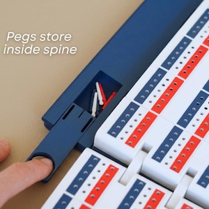 May include: A cribbage board with a blue spine and white playing surface, featuring red and blue scoring tracks. The text "Pegs store inside spine" is visible. The board includes a storage compartment for pegs.