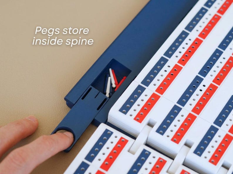 May include: A cribbage board with a blue spine and white playing surface, featuring red and blue scoring tracks. The text "Pegs store inside spine" is visible. The board includes a storage compartment for pegs.