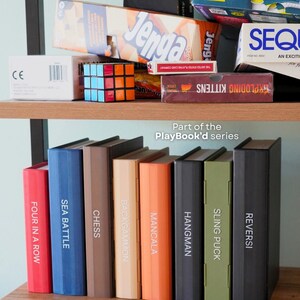 May include: A wooden shelf is filled with board games and books. Games include "Jenga," "Exploding Kittens," and a Rubik's Cube. Books from the "PlayBook'd series" are visible, with titles like "Four in a Row," "Sea Battle," and "Chess."
