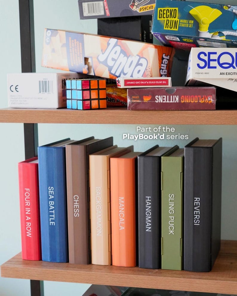 May include: A wooden shelf is filled with board games and books. Games include "Jenga," "Exploding Kittens," and a Rubik's Cube. Books from the "PlayBook'd series" are visible, with titles like "Four in a Row," "Sea Battle," and "Chess."