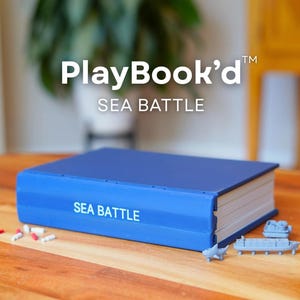 May include: A blue game box labeled "PlayBook'd SEA BATTLE" with miniature ship game pieces. The game box is rectangular and resembles a book. Small red and white game pieces are scattered on the wooden surface.