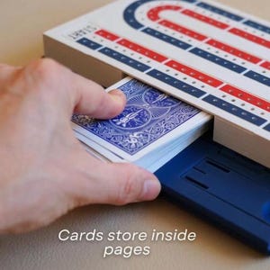 May include: A cribbage board with red, white, and blue detailing, and a deck of playing cards. The text "Cards store inside pages" is visible. The board has a blue storage compartment.