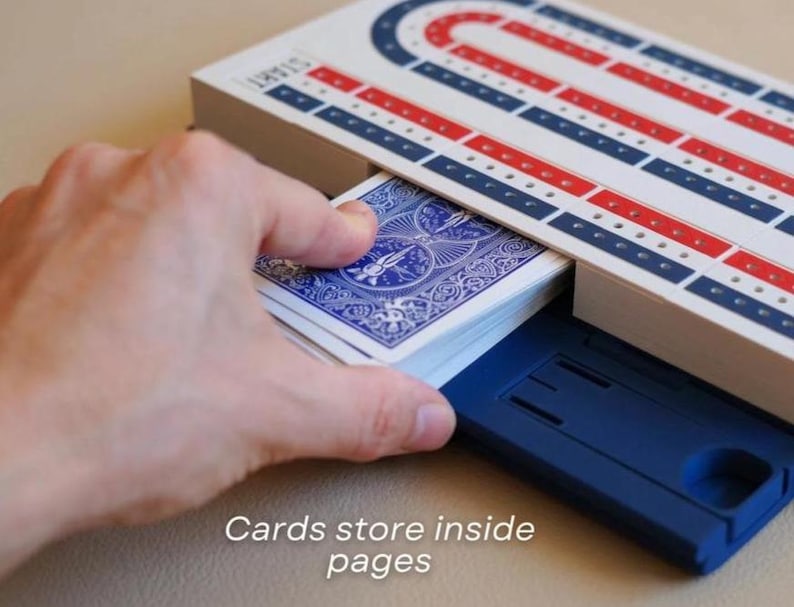 May include: A cribbage board with red, white, and blue detailing, and a deck of playing cards. The text "Cards store inside pages" is visible. The board has a blue storage compartment.