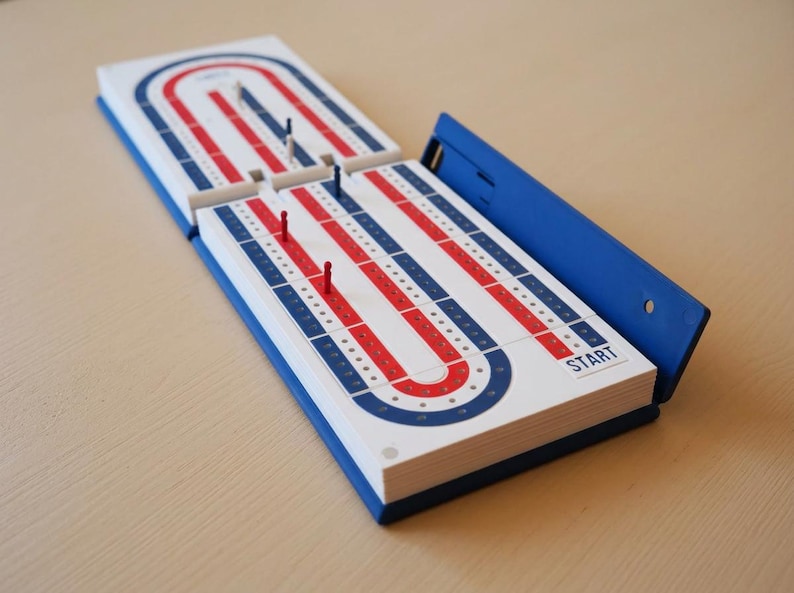 May include: A cribbage board game in blue, white, and red. The open board shows the playing surface with tracks and peg holes. The word "START" is visible. Several pegs are in place, ready for the game.