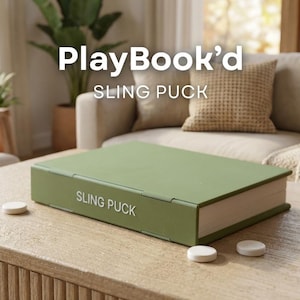 Puede incluir: Un juego de Sling Puck verde con diseño de libro, con el texto "PlayBook'd" y "SLING PUCK" visibles. El juego incluye discos blancos sobre una mesa de madera. El fondo presenta un sofá y plantas.