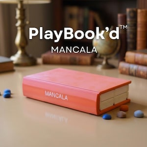 Puede incluir: Un juego de Mancala naranja con diseño de libro, con el texto "PlayBook'd" y "MANCALA". Varias piezas de juego azules, marrones y naranjas están dispersas alrededor del juego. El fondo incluye libros y una lámpara de escritorio.