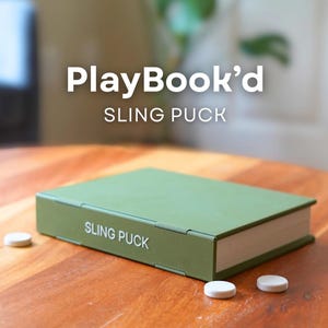 May include: A green rectangular game box labeled "SLING PUCK" with three white pucks on a wooden table. The text "PlayBook'd" is above the game box.