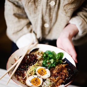 May include: A bowl of ramen with noodles, greens, and two halves of a boiled egg with sesame seeds. The bowl is held by a person wearing a tan jacket. Chopsticks rest on the bowl.