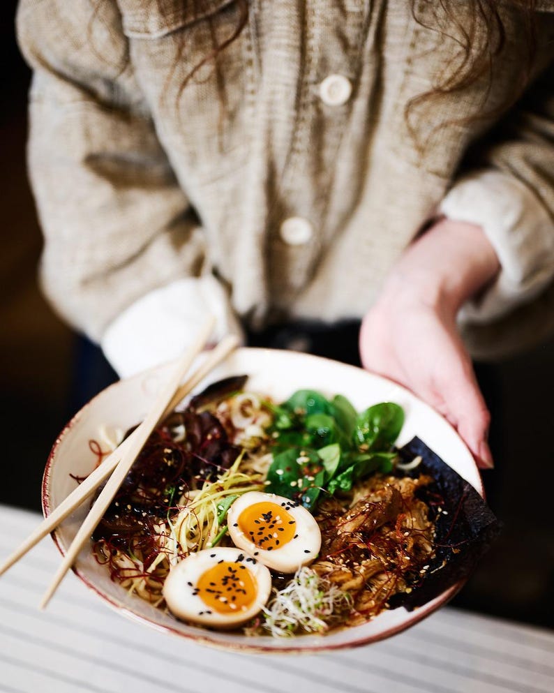 May include: A bowl of ramen with noodles, greens, and two halves of a boiled egg with sesame seeds. The bowl is held by a person wearing a tan jacket. Chopsticks rest on the bowl.