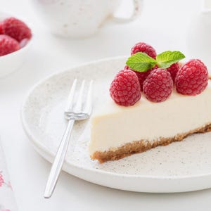 K&ouml;nnte beinhalten: Ein St&uuml;ck K&auml;sekuchen mit frischen Himbeeren und Minzbl&auml;ttern auf einem gesprenkelten wei&szlig;en Teller. Eine Sch&uuml;ssel mit Himbeeren, eine Gabel und eine Blumenserviette sind ebenfalls zu sehen.