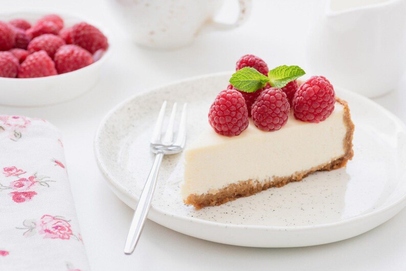 K&ouml;nnte beinhalten: Ein St&uuml;ck K&auml;sekuchen mit frischen Himbeeren und Minzbl&auml;ttern auf einem gesprenkelten wei&szlig;en Teller. Eine Sch&uuml;ssel mit Himbeeren, eine Gabel und eine Blumenserviette sind ebenfalls zu sehen.