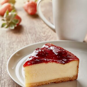 K&ouml;nnte beinhalten: Ein St&uuml;ck K&auml;sekuchen mit gl&auml;nzendem rotem Belag auf einem wei&szlig;en Teller. Eine wei&szlig;e Tasse Tee und frische Erdbeeren befinden sich im Hintergrund. Das Dessert befindet sich auf einer Holzoberfl&auml;che.