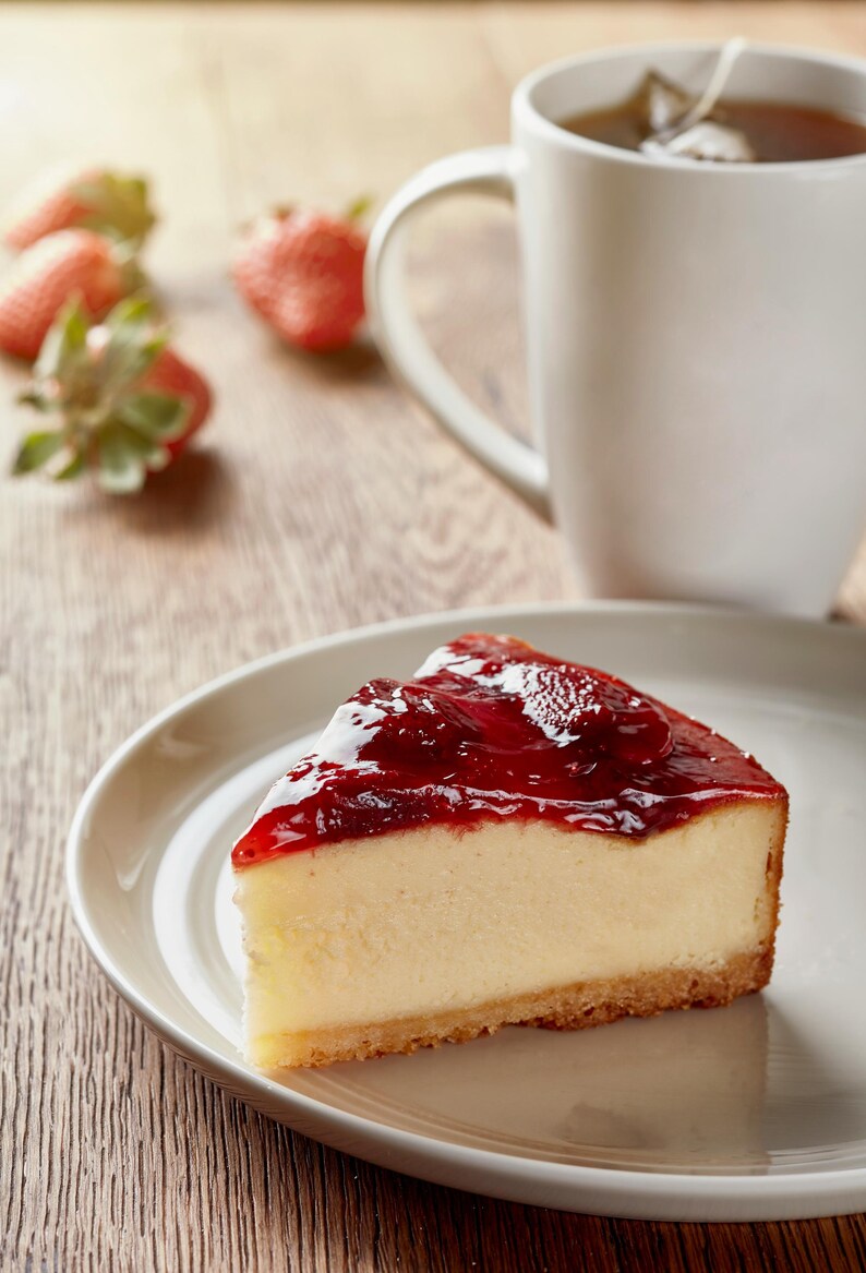 K&ouml;nnte beinhalten: Ein St&uuml;ck K&auml;sekuchen mit gl&auml;nzendem rotem Belag auf einem wei&szlig;en Teller. Eine wei&szlig;e Tasse Tee und frische Erdbeeren befinden sich im Hintergrund. Das Dessert befindet sich auf einer Holzoberfl&auml;che.