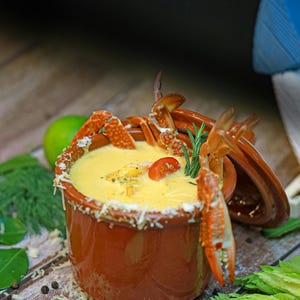 May include: A bowl of creamy yellow soup with crab claws and a tomato garnish. The rim of the bowl is decorated with grated cheese. Fresh herbs and spices are scattered around the bowl on a wooden surface.