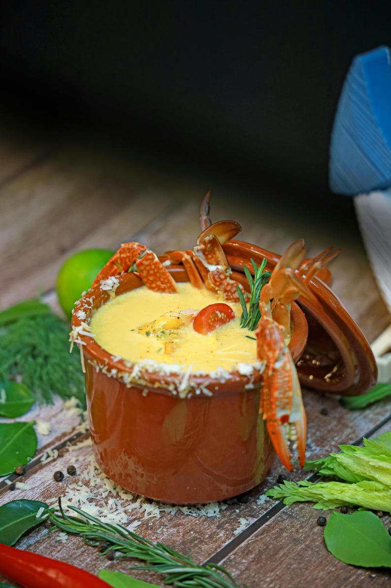 May include: A bowl of creamy yellow soup with crab claws and a tomato garnish. The rim of the bowl is decorated with grated cheese. Fresh herbs and spices are scattered around the bowl on a wooden surface.