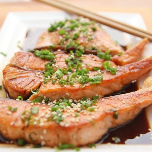 May include: Grilled salmon fillets on a white rectangular plate, garnished with sesame seeds and chopped green onions. The salmon is served with a dark sauce, and chopsticks are placed on the side.