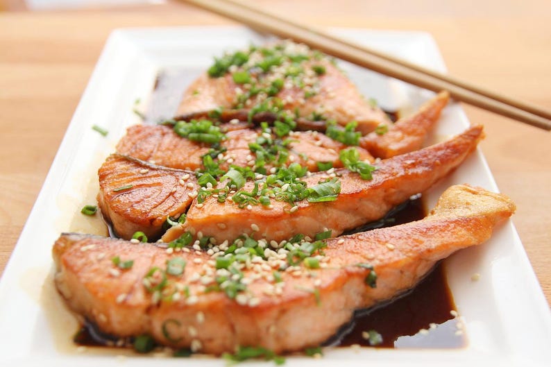 May include: Grilled salmon fillets on a white rectangular plate, garnished with sesame seeds and chopped green onions. The salmon is served with a dark sauce, and chopsticks are placed on the side.