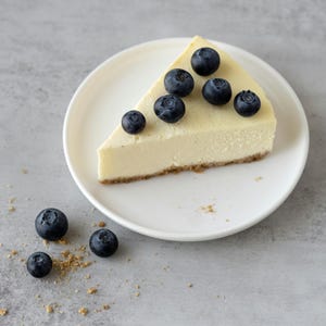 K&ouml;nnte beinhalten: Ein St&uuml;ck K&auml;sekuchen auf einem wei&szlig;en Teller, garniert mit frischen Blaubeeren. Der K&auml;sekuchen hat eine goldgelbe F&uuml;llung und einen Keksboden. Blaubeeren und Kr&uuml;mel sind auf der grauen Oberfl&auml;che verstreut.