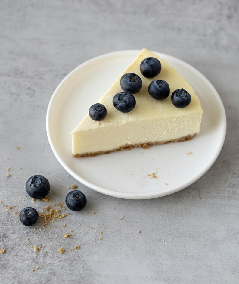K&ouml;nnte beinhalten: Ein St&uuml;ck K&auml;sekuchen auf einem wei&szlig;en Teller, garniert mit frischen Blaubeeren. Der K&auml;sekuchen hat eine goldgelbe F&uuml;llung und einen Keksboden. Blaubeeren und Kr&uuml;mel sind auf der grauen Oberfl&auml;che verstreut.