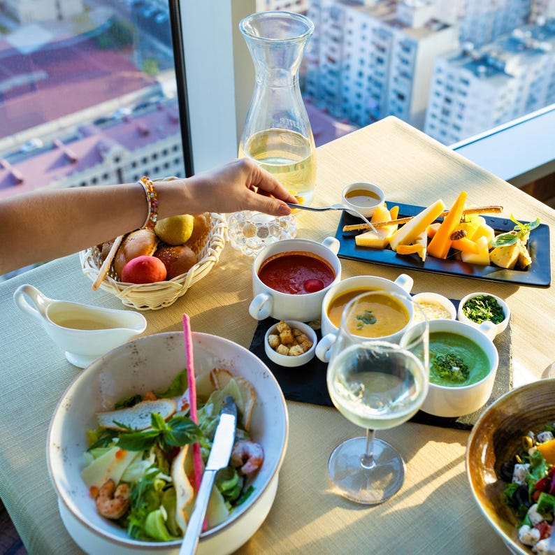 May include: A table setting with various dishes, including soup, salad, cheese, and bread. A clear glass carafe and a glass of white wine are also present. The scene is set near a window with a city view.