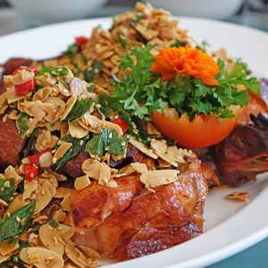 May include: A plate of roasted chicken with a crispy, golden-brown skin, topped with a mix of toasted almonds, herbs, and red chili peppers. Garnished with a tomato and an orange flower. The dish is served on a white plate.