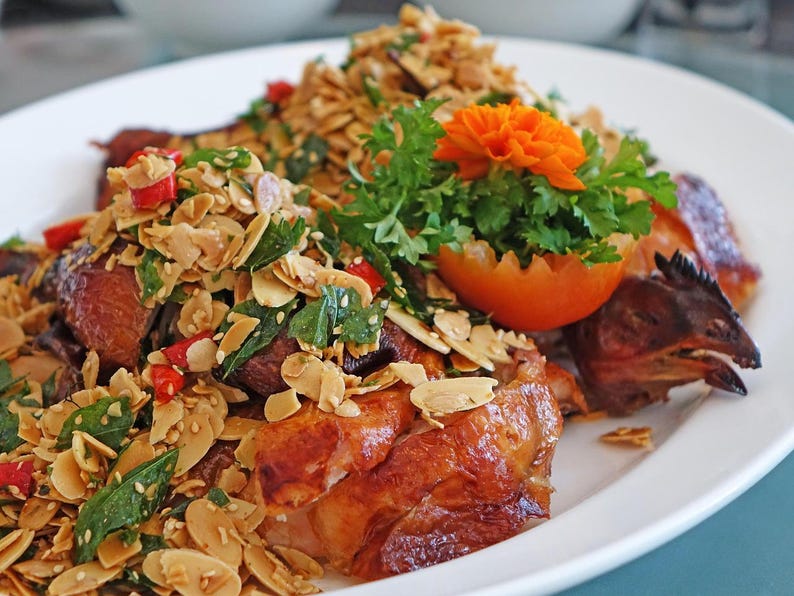 May include: A plate of roasted chicken with a crispy, golden-brown skin, topped with a mix of toasted almonds, herbs, and red chili peppers. Garnished with a tomato and an orange flower. The dish is served on a white plate.