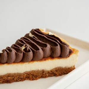 K&ouml;nnte beinhalten: Ein St&uuml;ck K&auml;sekuchen mit Keksboden, cremiger wei&szlig;er F&uuml;llung und dekorativem dunklem Schokoladenfrosting. Goldblatt akzentuiert die Oberseite. Das Dessert liegt auf einem wei&szlig;en rechteckigen Teller.