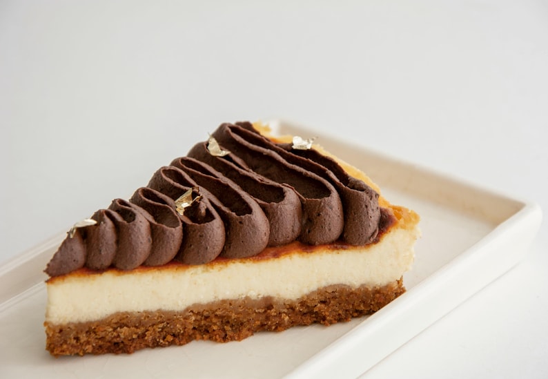 K&ouml;nnte beinhalten: Ein St&uuml;ck K&auml;sekuchen mit Keksboden, cremiger wei&szlig;er F&uuml;llung und dekorativem dunklem Schokoladenfrosting. Goldblatt akzentuiert die Oberseite. Das Dessert liegt auf einem wei&szlig;en rechteckigen Teller.