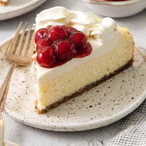 K&ouml;nnte beinhalten: Ein St&uuml;ck K&auml;sekuchen mit Keksboden, garniert mit Schlagsahne und glasierten Kirschen. Das Dessert befindet sich auf einem gesprenkelten Teller neben einer Vintage-Gabel. Der K&auml;sekuchen hat eine cremige, hellgelbe Farbe.