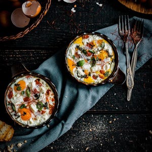 May include: Overhead shot of two small cast iron skillets filled with baked eggs, tomatoes, and herbs. A basket of brown eggs and bread are nearby. The food is on a dark wooden surface with a blue cloth and two forks.