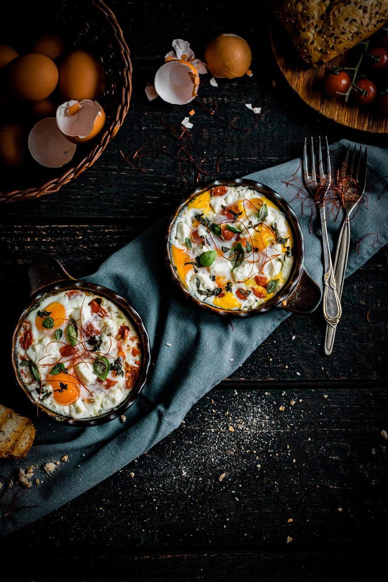 May include: Overhead shot of two small cast iron skillets filled with baked eggs, tomatoes, and herbs. A basket of brown eggs and bread are nearby. The food is on a dark wooden surface with a blue cloth and two forks.