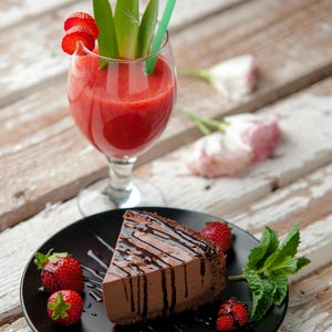 K&ouml;nnte beinhalten: Ein St&uuml;ck Schokoladenkuchen mit Schokoladenguss auf einem schwarzen Teller, garniert mit frischen Erdbeeren und Minze. Ein Glas roter Smoothie mit gr&uuml;nem Strohhalm und Erdbeer-Garnitur. Auf einer Holzoberfl&auml;che.