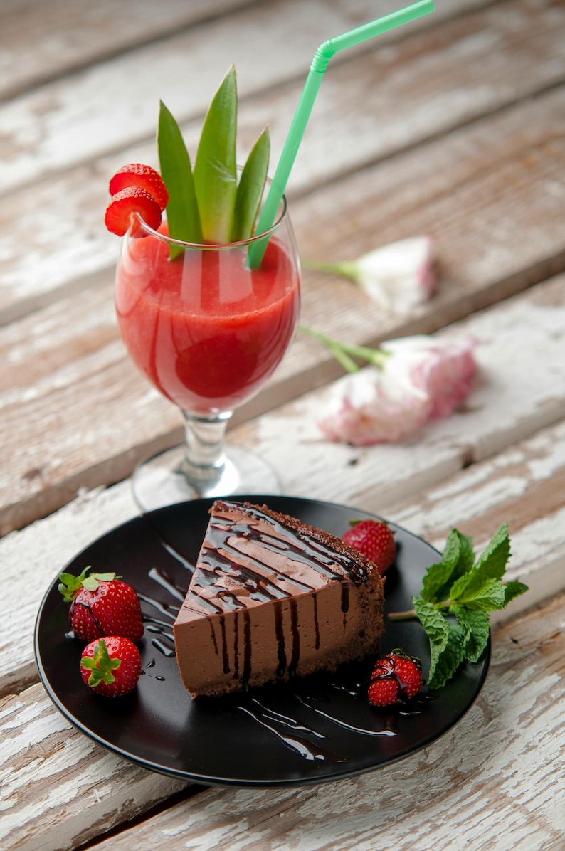 K&ouml;nnte beinhalten: Ein St&uuml;ck Schokoladenkuchen mit Schokoladenguss auf einem schwarzen Teller, garniert mit frischen Erdbeeren und Minze. Ein Glas roter Smoothie mit gr&uuml;nem Strohhalm und Erdbeer-Garnitur. Auf einer Holzoberfl&auml;che.