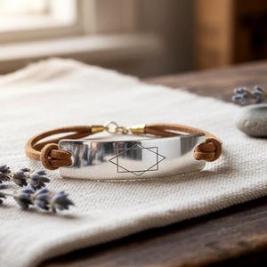 Könnte beinhalten: Ein silbernes Armband mit geometrischem Design und braunem Lederband. Das Armband wird auf einer weißen Leinenoberfläche präsentiert, mit Lavendelzweigen und einem kleinen Stein im Hintergrund.